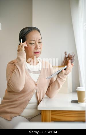 Ritratto di una donna asiatica pensata e anziana in abiti comodi utilizzando il suo tablet digitale, lavorando alla sua attività mentre si rilassa in una caffetteria. Foto Stock