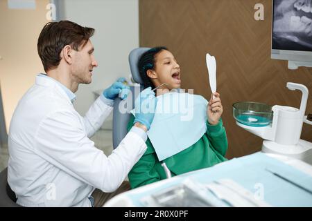 Vista laterale ritratto di ragazza adolescente nera che guarda in specchio durante il controllo dentale in clinica Foto Stock