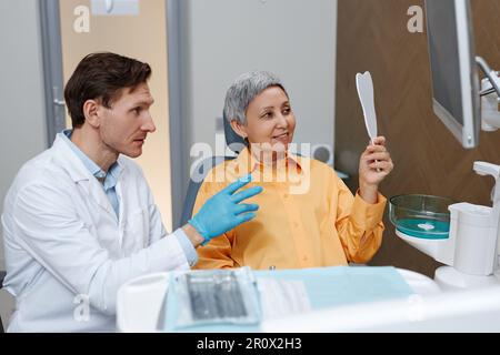 Vista laterale ritratto di donna anziana che guarda in specchio alla clinica dentale e sorridente Foto Stock