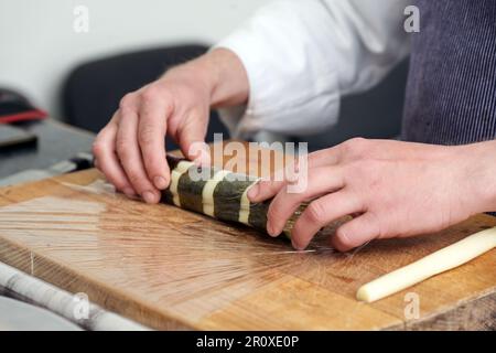 Mani di uno chef che arrotola asparagi bianchi in foglie di alghe nori su un tavolo da cucina, preparazione per una cena gourmet vegetariana, focus selezionato Foto Stock