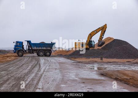 Ust-Luga, Leningrado oblast, Russia - 16 novembre 2021: L'escavatore Caterpillar carica la ghiaia nel dumper. Strada a terra bagnata in primo piano. Foto Stock