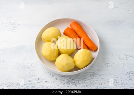 Piatto bianco con verdure sbucciate, patate e carote su sfondo azzurro, vista dall'alto. Fase di cottura dell'insalata di verdure Foto Stock