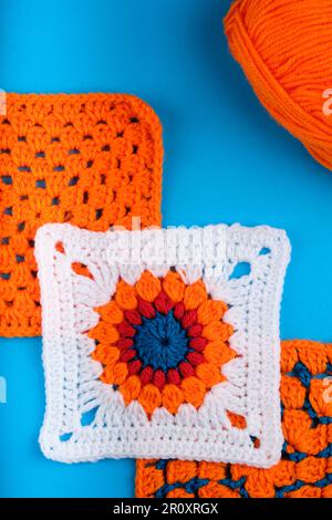 White Granny Square con motivo arancione centrale, rosso e blu. Due quadrati di aranciata e una sfera di filo aranciata dietro tutto su Kingfisher Blue. Foto Stock