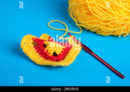 Crochet Granny Square in giallo e rosso con palla gialla di filo e gancio a crochet rosso scuro su sfondo blu Kingfisher. Foto Stock