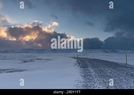 Vista panoramica su un paesaggio innevato di lava, rocce e montagne con stretta strada sterrata in Islanda durante l'inverno con sole luminoso Foto Stock