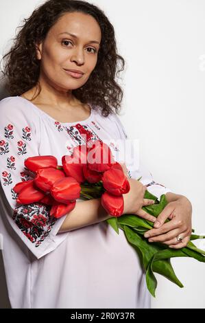 Multi etnico affascinante donna incinta in abito ricamato tradizionale, con bouquet di tulipani rossi, isolato su bianco Foto Stock