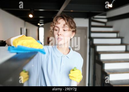 Giovane donna che indossa guanti protettivi con detergente per rimuovere la polvere dai mobili durante i lavori domestici professionali Foto Stock