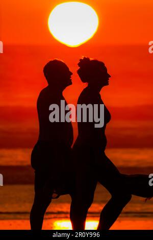 Uomo e donna che camminano con silhouette al tramonto sulla spiaggia di Guiones, dove molti si riuniscono per guardare il sole tramontare. Playa Guiones, Nosara, Guanacaste, Costa Rica Foto Stock