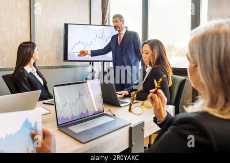 I colleghi multietnici si sono concentrati in abiti formali che si riuniscono al tavolo con i computer portatili mentre si brainstorming e si analizzano i grafici durante le riunioni finanziarie Foto Stock