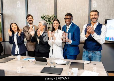 Gruppo di colleghi multietnici in formale indossare sorridente e lappare le mani durante la riunione in ufficio moderno Foto Stock