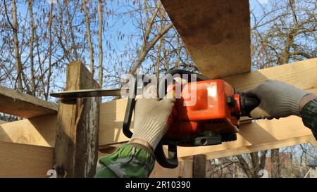 tagliando la parte in eccesso della tavola di una struttura in legno di un edificio esterno utilizzando una motosega, un costruttore rurale rifinisce il bordo della tavola Foto Stock