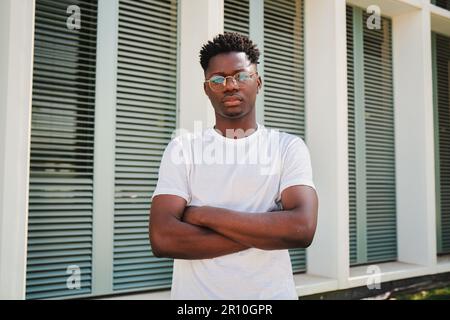 Ritratto di giovane afro-americano serio ragazzo fiducioso guardando la macchina fotografica con braccia incrociate in piedi all'esterno. Bell'uomo studente universitario che si pone con un atteggiamento calmo e penoso presso il campus universitario. Foto di alta qualità Foto Stock