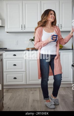 Che cosa dovrei cucinare per pranzo. una bella giovane donna che ha il caffè mentre si è in piedi accanto al frigorifero in cucina a casa. Foto Stock