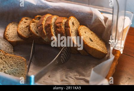 Pane integrale a fette in cesto su un tavolo di legno rustico marrone scuro, luce naturale dalla finestra. Foto Stock