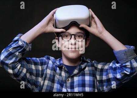 Un ragazzo caucasico di 12 anni che indossa occhiali e una camicia a scacchi solleva il visore VR, appoggiandolo sulla testa, mentre guarda la telecamera e sorride. Foto Stock