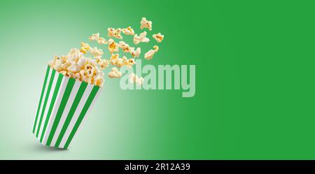 Popcorn che vola fuori dalla scatola di carta verde a strisce bianche isolato su sfondo verde con spazio di copia. Spruzzi, levitazione di grani di popcorn. Foto Stock