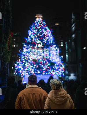 Un gruppo di persone che ammirano un vivace albero di Natale in città illuminato da luci colorate Foto Stock