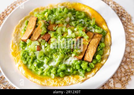 Omelette all'uovo con purè di piselli freschi verdi e gambo di sedano e tofu affumicato su piatto bianco, su tampone di bambù, primo piano. Foto Stock