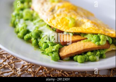 Omelette all'uovo con purè di piselli freschi, gambo di sedano e tofu affumicato su piatto bianco su tampone di bambù, primo piano. Delizioso pasto vegetariano. Foto Stock