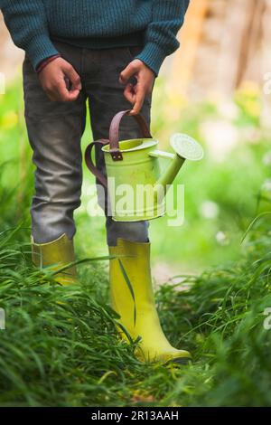 Una scena giocosa si svolge come le gambe di un ragazzo, rivestito di stivali da pioggia, brandisci un'annaffiatura, abbracciando la gioia di spruzzare e annaffiare le piante. Foto Stock