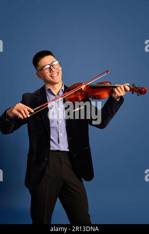 Ritratto con bei musicisti giovani, violinista che indossa un abito elegante e occhiali che suonano sul violino e sorridono su sfondo blu dello studio Foto Stock