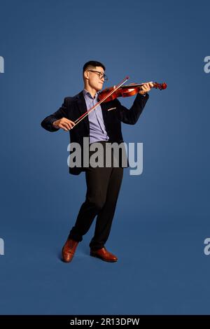 Ritratto con musicisti giovani e professionisti, violinista che indossa un costume classico con fidle bow e suona sul violino e sorride sul blu Foto Stock
