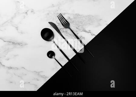 Vista dall'alto delle posate nere su marmo bianco e sfondo nero. Piano di impostazione creativa tavolo piatto. Forchetta, coltello, cucchiaio e cucchiaio da dessert. Spazio di copia. Foto Stock
