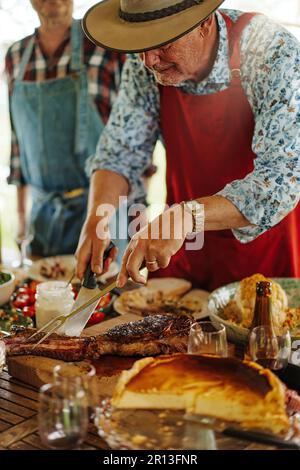 Un uomo di 65 anni che indossa un cappello di paglia e un grembiule taglia abilmente carne alla griglia con un coltello e una forchetta per barbecue. Sullo sfondo, persone irriconoscibili Foto Stock