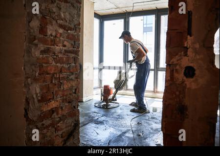 Lavoratore maschio che utilizza la macchina per la spianatura durante la spianatura del pavimento in appartamento in fase di ristrutturazione. Finitura di superfici cementate con smerigliatrice per pavimenti in camera con grande finestra. Foto Stock