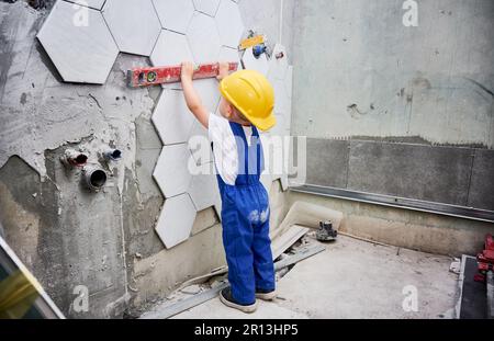 Piccolo ragazzo che controlla la superficie della parete con la livella a bolla d'aria in appartamento. Bambino in tute da lavoro utilizzando strumenti di livello durante il lavoro di ristrutturazione a casa. Foto Stock