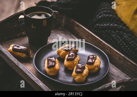 Mini eclair con cioccolato fondente e un caffè su un vassoio di legno, specialità di pasticceria francese con crema pasticcera Foto Stock