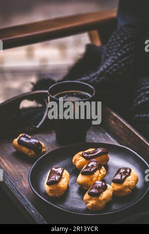 Vassoio di legno con mini eclairs e caffè su un vassoio di legno su una poltrona di fronte alla finestra, coffee break, verticale Foto Stock