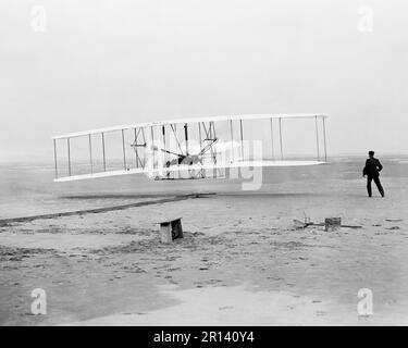 Primo volo, 120 piedi in 12 secondi; Kitty Hawk, Carolina del Nord. La fotografia mostra Orville Wright ai comandi della macchina volante, giacendo prono sull'ala inferiore con fianchi nella culla che azionava il meccanismo di orditura delle ali. Wilbur Wright che corre accanto per bilanciare la macchina, ha appena rilasciato la sua presa sul diritto in avanti dell'ala destra. Dietro la macchina sono visibili il binario di partenza, il poggialettone, una scatola delle bobine e altri elementi necessari per la preparazione del volo. Fotografia di John T. Daniels Foto Stock