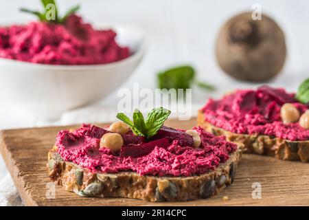 Asse di legno di fette di pane con hummus di barbabietola su uno sfondo di legno bianco Foto Stock