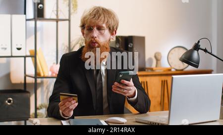 Bearded businessman programmatore software sviluppatore di lavoro fare acquisti online pagamento con carta di credito smartphone presso la moderna scrivania home Office con computer portatile. Giovane uomo d'affari freelance Foto Stock