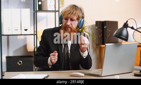 Felice rilassato giovane uomo d'affari gioioso che lavora sul computer portatile in casa ufficio indossando cuffie ascoltare musica da discoteca preferita energetica su smartphone. Uomo freelance che si rilassa, facendo una pausa Foto Stock