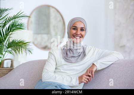 Ritratto di giovane donna d'affari di origine orientale in hijab seduto sul divano a casa e guardando la macchina fotografica con fiducia e sorridente. Foto Stock