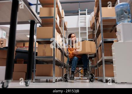 Responsabile del magazzino in sedia a rotelle che controlla la qualità dell'imballaggio dei pacchi nel magazzino. Operatore logistico che trasporta la scatola di cartone mentre fa la gestione dell'inventario delle scorte in deposito accessibile ai disabili Foto Stock