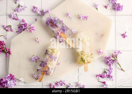 Tavola con bellissimi popsicles floreali e fiori di lilla su sfondo piastrelle chiaro Foto Stock