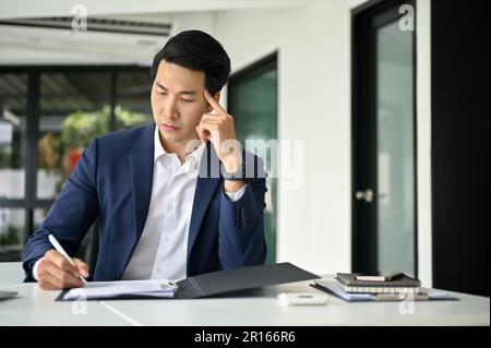 Uomo d'affari asiatico o banchiere maschile sottolineato e premuroso millenario in tuta professionale di affari che esamina i rapporti finanziari o di revisione del business d Foto Stock