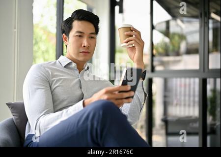 Un uomo d'affari asiatico o un banchiere maschile intelligente e bello che controlla le e-mail aziendali sul suo smartphone mentre sorseggia un caffè e riposa nei suoi offi Foto Stock