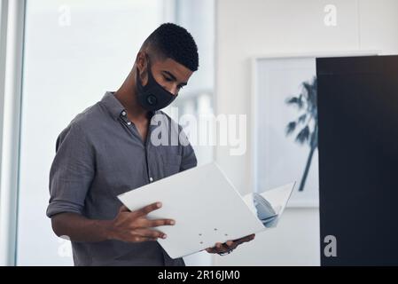 Sfogliare i file aziendali. un giovane uomo d'affari che indossa una maschera facciale mentre passa attraverso una cartella in un ufficio. Foto Stock