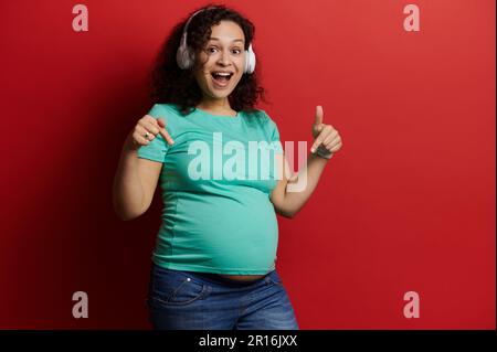Felice donna incinta con cuffie wireless, ascolta musica, punta il dito sul ventre, rosso isolato Foto Stock