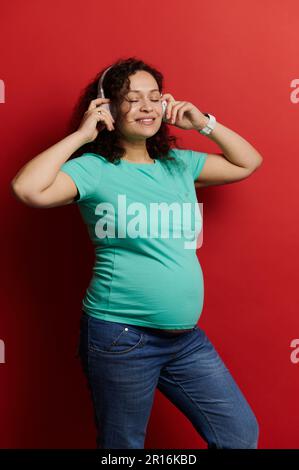 Ritratto di una bella donna incinta godendo di musica rilassante, indossando cuffie wireless, isolato su sfondo rosso Foto Stock