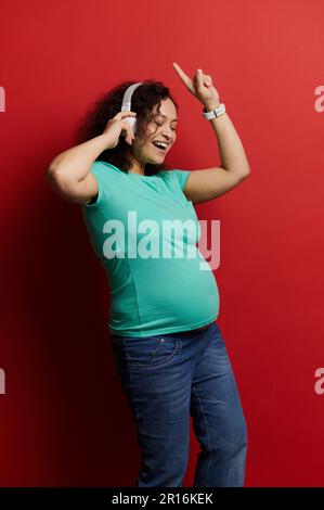 Donna incinta positiva che indossa cuffie wireless, divertirsi, ballare, ascoltare musica, isolato su sfondo rosso Foto Stock