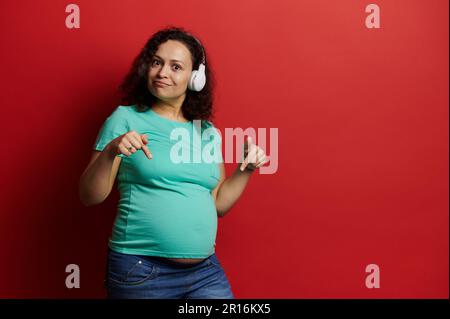 Affascinante donna adulta incinta che mostra il ventre alla fotocamera, ascoltando musica con cuffie wireless su sfondo rosso Foto Stock