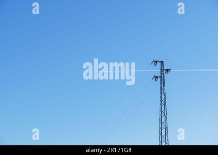 Un aereo passa lasciando la sua scia dietro una torre ad alta tensione, sullo sfondo un cielo azzurro chiaro Foto Stock