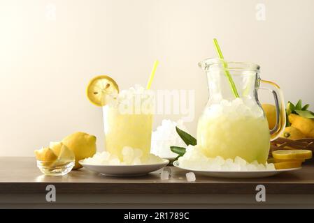 Limonata con ghiaccio in una brocca e bicchiere su una panca di legno da cucina con frutta e ghiaccio tritato con fondo isolato. Vista frontale. Composizione orizzontale Foto Stock