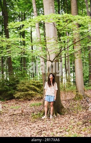 ragazza in pantaloncini si alza sotto l'albero guardando il verde in uno stato di riposo Foto Stock