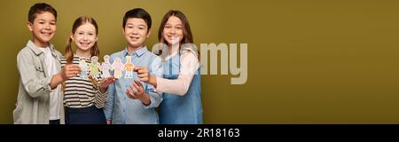 Amici premianti interrazziali felici in vestiti casuali che tengono i caratteri disegnati del mestiere della carta e che guardano la macchina fotografica durante la celebrazione di giorno di protezione del bambino Foto Stock